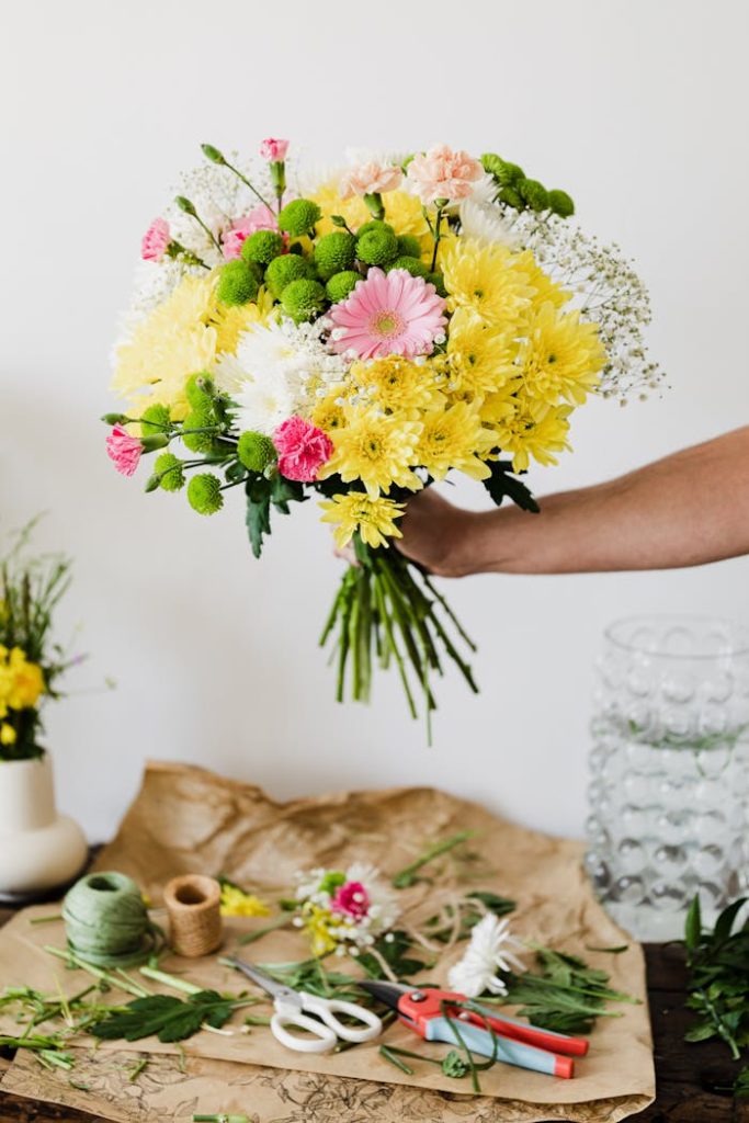 Crop faceless florist composing chrysanthemum bouquet in florist workshop at messy table covered with scattered leaves and bits of stems